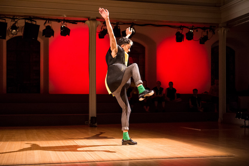 Caleb Teicher wears a black and yellow tunic, grey slacks, green socks and tap shoes. Both of his arms are stretched to the ceiling while one leg is hiked up in the air.